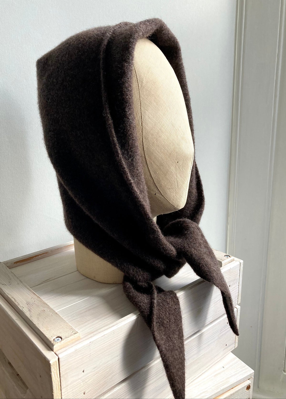 A dark brown wool bonnet or headscarf with long ties, displayed on a mannequin head resting on a light-colored wooden crate against a white wall.