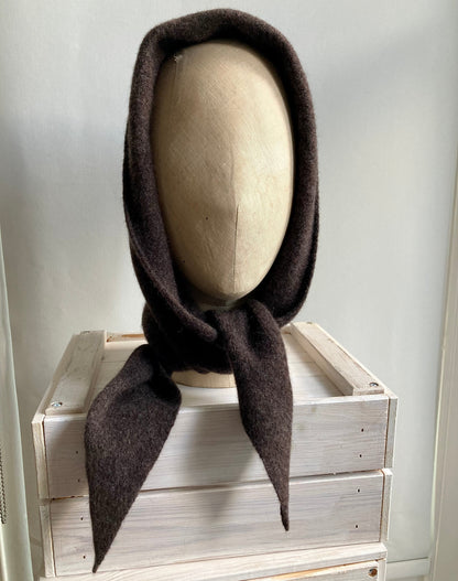 A dark brown wool bonnet with long tails, displayed on a mannequin head resting on a light-colored wooden crate.