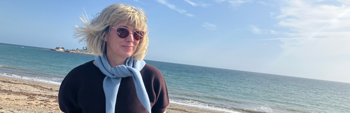 A woman with blonde hair and sunglasses stands on a beach wearing a brown top and a light blue neckerchief scarf, with the sea and blue sky in the background.