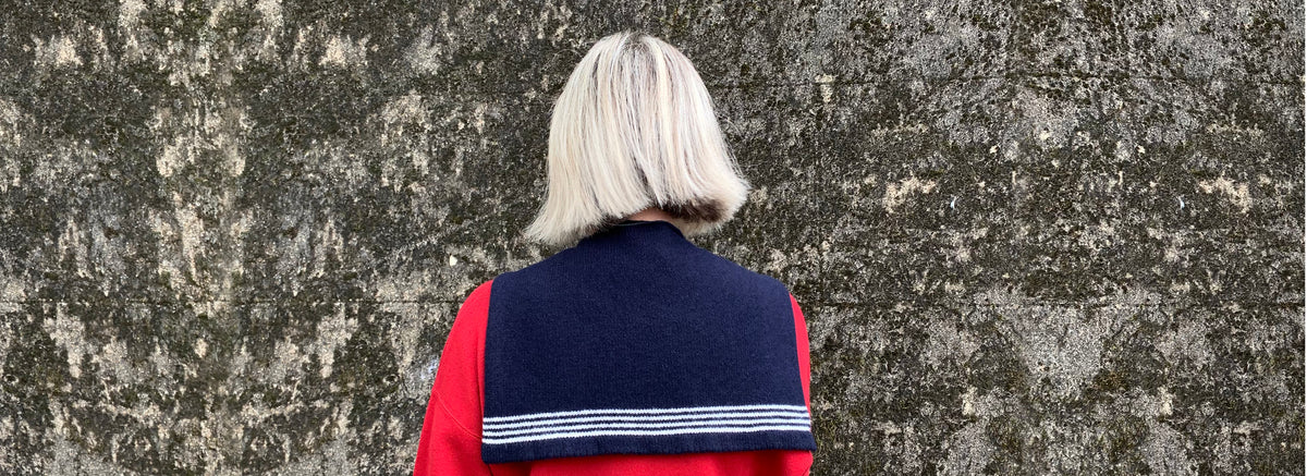 A person viewed from behind wearing a navy blue sailor collar scarf with white stripes, over a red top, standing against a textured grey stone wall.