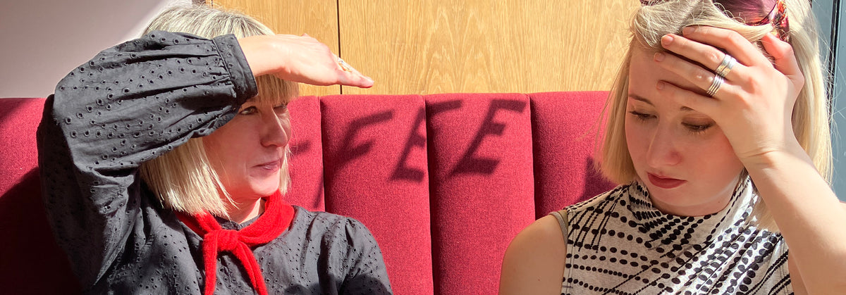 Two women sitting side-by-side in a red upholstered booth, one wearing a red knitted neckerchief bandana.