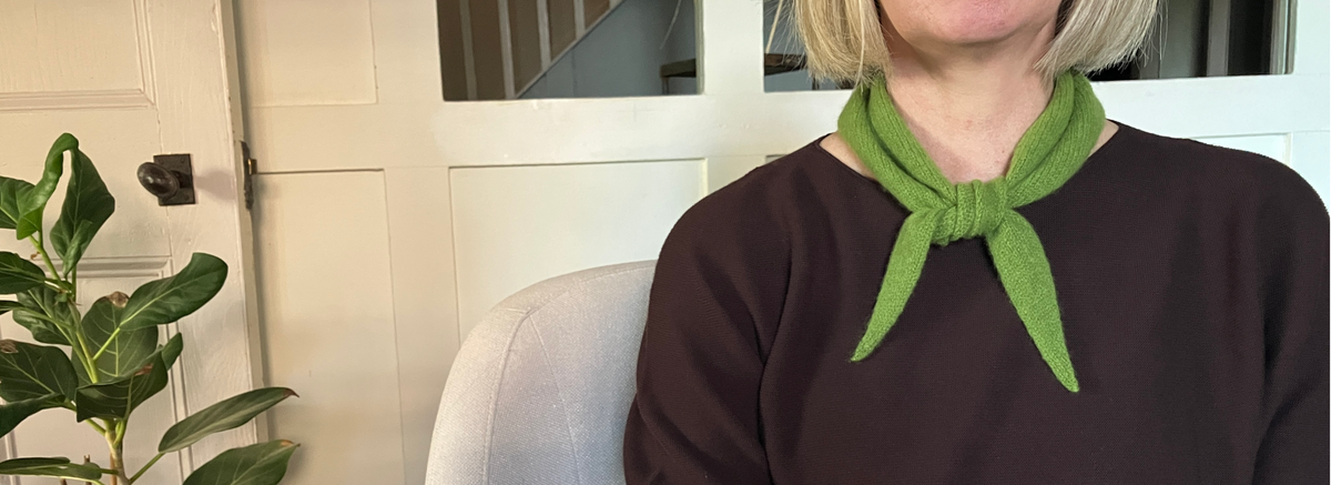 A person wearing a dark brown top and a bright green neckerchief scarf, seated indoors in front of a white panelled door with a houseplant visible on the left.