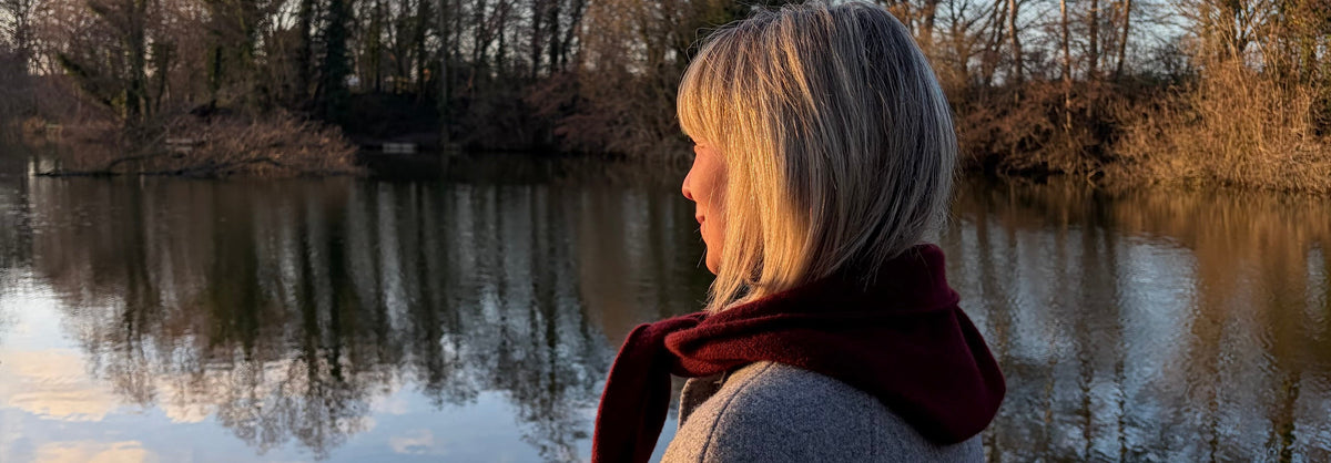 A woman with blonde hair is shown in profile, wearing a red brown hooded scarf and a light grey coat, standing by a calm lake with bare trees in the background during golden hour.